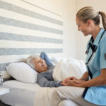 Lächelnde Krankenschwester spricht mit ihrer Patientin, einer älteren Frau, die zuhause im Bett liegt